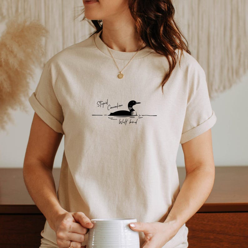 Pu&ograve; includere: Una t-shirt beige a maniche corte con una grafica nera di un colimbo che nuota sull'acqua. Il testo "Stupid Canadian Wolf bird" &egrave; stampato in un carattere corsivo nero sopra e sotto l'illustrazione al centro del petto.