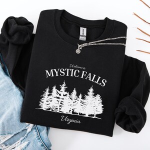 Könnte beinhalten: Ein schwarzes Langarm-Sweatshirt mit weißem Siebdrucktext, der Welcome to MYSTIC FALLS Virginia lautet, über einer grafischen Illustration eines dichten Kiefernwaldes. Das Kleidungsstück hat einen Rundhalsausschnitt und ein Gildan-Markenetikett.