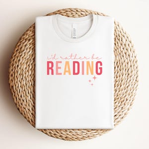 Op de afbeelding: Wit T-shirt met de tekst "i'd rather be READING" in roze, oranje en geel met kleine roze sterren.