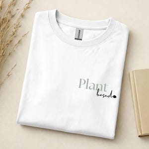 May include: A white crew neck t-shirt featuring the text Plant based printed on the front. The word Plant is in a large grey serif font, while the word based is in a smaller black script font with a small leaf icon at the end of the text.