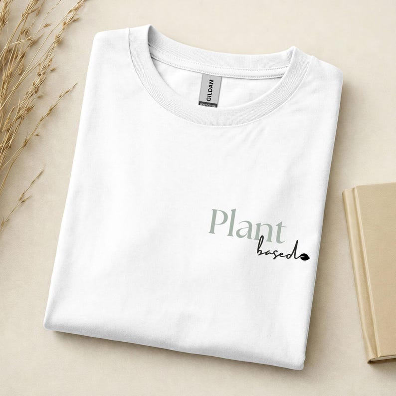 May include: A white crew neck t-shirt featuring the text Plant based printed on the front. The word Plant is in a large grey serif font, while the word based is in a smaller black script font with a small leaf icon at the end of the text.