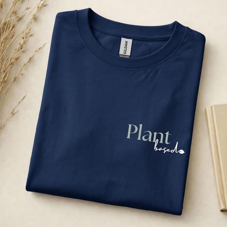 May include: A navy blue short-sleeved t-shirt featuring the text Plant based printed in a combination of serif and script fonts on the lower right side. The shirt has a crew neckline and a Gildan brand label visible at the inner collar area.