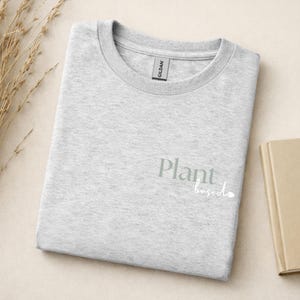 May include: A light gray crew neck t-shirt with a Gildan brand tag at the collar. The front of the shirt features the text Plant based printed in a sage green serif font for the word Plant and a white cursive script for the word based.