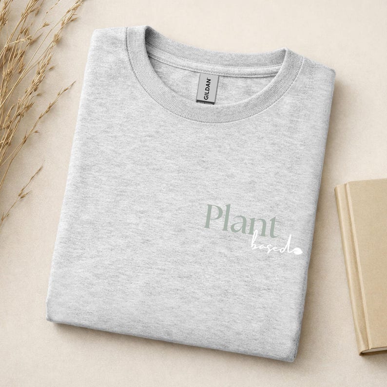 May include: A light gray crew neck t-shirt with a Gildan brand tag at the collar. The front of the shirt features the text Plant based printed in a sage green serif font for the word Plant and a white cursive script for the word based.
