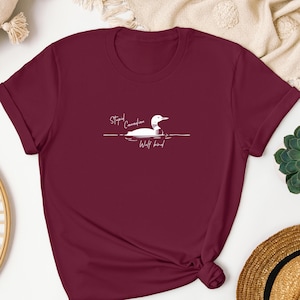 Pu&ograve; includere: Una t-shirt a maniche corte color bordeaux con una grafica bianca che raffigura un colimbo che nuota sull'acqua. Il testo "Stupid Canadian Wolf bird" &egrave; stampato in un carattere corsivo bianco sopra e sotto l'illustrazione dell'uccello.