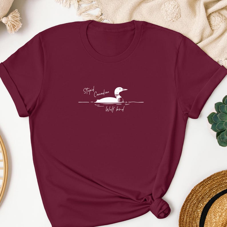 Pu&ograve; includere: Una t-shirt a maniche corte color bordeaux con una grafica bianca che raffigura un colimbo che nuota sull'acqua. Il testo "Stupid Canadian Wolf bird" &egrave; stampato in un carattere corsivo bianco sopra e sotto l'illustrazione dell'uccello.