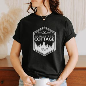 Könnte beinhalten: Schwarzes T-Shirt mit weißem Aufdruck. Der Aufdruck ist ein sechseckiges Design mit dem Text "I'M COMING TO THE COTTAGE CANADA" und gekreuzten Hockeyschlägern. Das T-Shirt hat einen Rundhalsausschnitt.