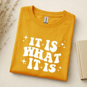 Könnte beinhalten: Ein senfgelbes T-Shirt mit Rundhalsausschnitt und dem weißen Schriftzug "IT IS WHAT IT IS" in einer fetten, wellenförmigen Schrift. Das Design ist mit mehreren kleinen, vierzackigen Sternen um den Text herum auf der Vorderseite bedruckt.