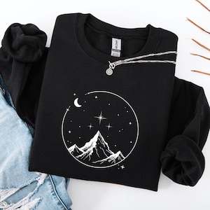 Könnte beinhalten: Schwarzes Sweatshirt mit Rundhalsausschnitt und weißer Grafik eines Bergmassivs, Mond und Sterne in einem Kreis. Eine silberne Halskette mit einem Sonnenstrahl-Anhänger liegt auf dem Sweatshirt. Das Sweatshirt ist auf einer blauen Jeans gefaltet.