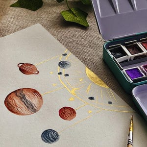 May include: Watercolor painting of a planetary system with brown, red, and blue planets connected by gold lines. A gold sun is in the background. A teal watercolor paint set and a paintbrush are in the frame.
