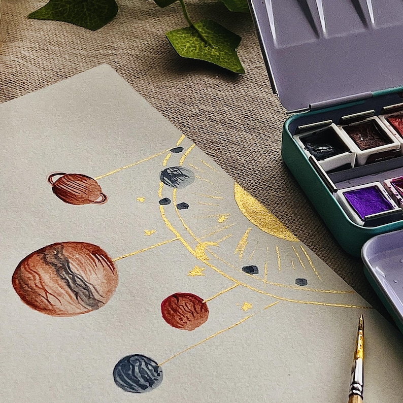 May include: Watercolor painting of a planetary system with brown, red, and blue planets connected by gold lines. A gold sun is in the background. A teal watercolor paint set and a paintbrush are in the frame.