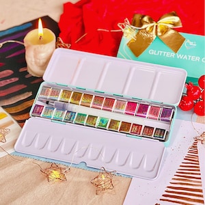 May include: An open, rectangular watercolor paint set with a light blue exterior and a white interior, filled with various colors. A brush and a small watercolor painting are visible. A lit candle and a card that says "Merry Christmas" are also in the image.