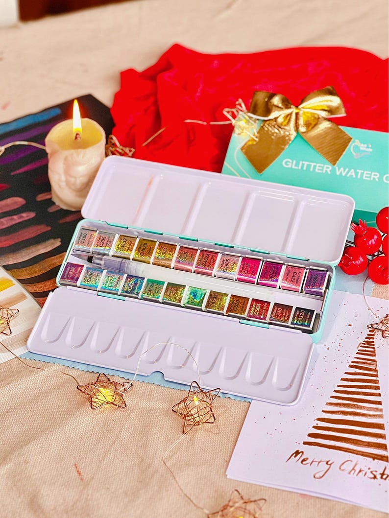 May include: An open, rectangular watercolor paint set with a light blue exterior and a white interior, filled with various colors. A brush and a small watercolor painting are visible. A lit candle and a card that says "Merry Christmas" are also in the image.
