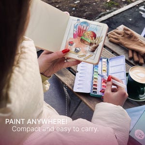 May include: A person is painting with watercolors in a sketchbook. The sketchbook is open to a drawing of a burger, fries, and drinks. A watercolor paint set and a cup of coffee are also visible. The text "PAINT ANYWHERE! Compact and easy to carry" is at the bottom.