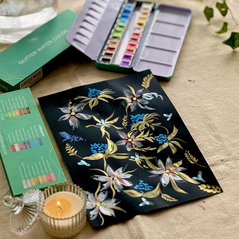 May include: A still-life composition featuring art supplies. A black paper with a floral design in gold, white, and blue is displayed alongside a green box of glitter watercolor paints, a metal paint palette, and a lit candle.