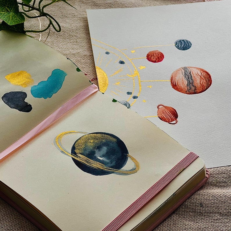 May include: Watercolor paintings of planets and a solar system. One painting features a dark blue planet with a gold ring. Another shows a solar system with planets in red, blue, and brown. The paintings are in a notebook.