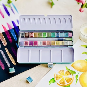 May include: A teal watercolor paint set with 24 color blocks in a metal tin. The set includes a water brush and is displayed open, revealing the various colors. Watercolor art supplies are visible in the background.