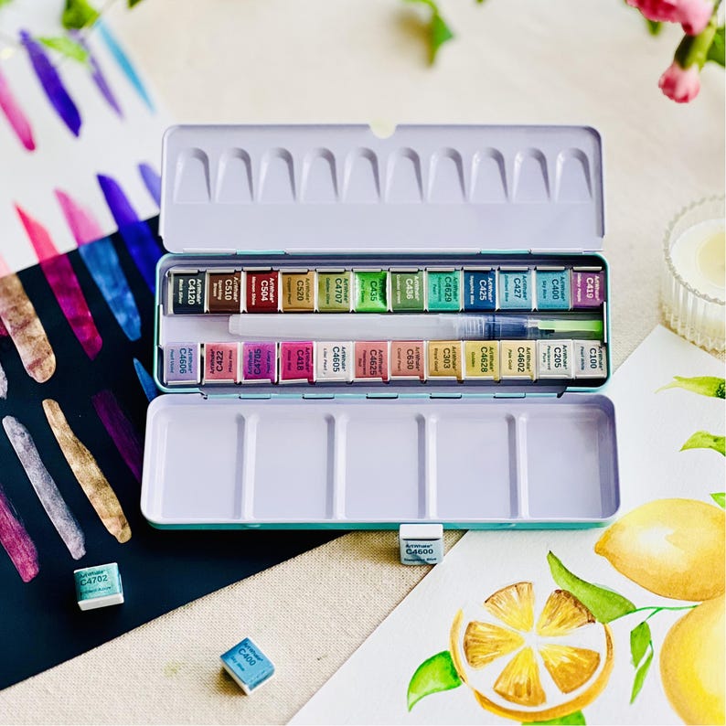 May include: A teal watercolor paint set with 24 color blocks in a metal tin. The set includes a water brush and is displayed open, revealing the various colors. Watercolor art supplies are visible in the background.