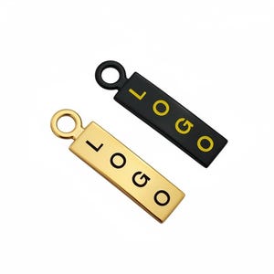 May include: Two rectangular metal charms, one gold and one black, each with the word "LOGO" in a contrasting color. The gold charm has a gold loop, and the black charm has a black loop. The charms are likely for jewelry or branding.