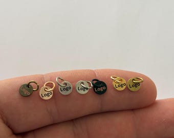 May include: A collection of six small, circular metal charms, each featuring a jump ring and the word Logo printed on the front. The charms are presented in various metallic finishes, including bronze, rose gold, silver, matte silver, black, and gold.