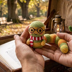 May include: A small, round, crocheted worm toy with a green and orange segmented body, wearing black wire-rimmed glasses and a red and white striped scarf. The toy has a smiling face with pink cheeks and is held in two hands above an open suitcase.