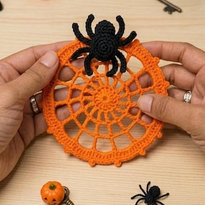 May include: A circular orange crocheted spider web coaster featuring a small black crocheted spider attached to the top. The web design consists of concentric rings connected by radiating spokes, creating a detailed, textured, and handmade appearance.