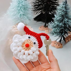 May include: A handmade crochet Santa Claus keyring. The keyring features a white beard and face, a red hat with a white pom-pom, and googly eyes. The keyring is holding a silver key. The background includes miniature Christmas trees.
