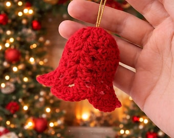 May include: A handmade, red crocheted bell Christmas ornament. The bell is held by a gold string and has a textured surface. The background features a Christmas tree with red ornaments and twinkling lights.