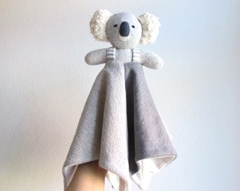 May include: A grey and white koala soft toy with a white and grey blanket attached. The koala is wearing a grey and white striped bib.