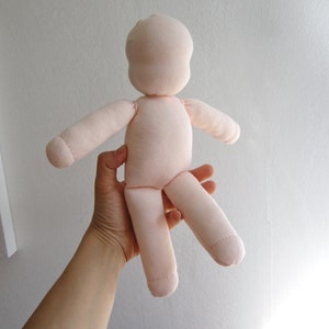 May include: A plain, light pink fabric doll with no facial features. The doll is held by a hand in the air.