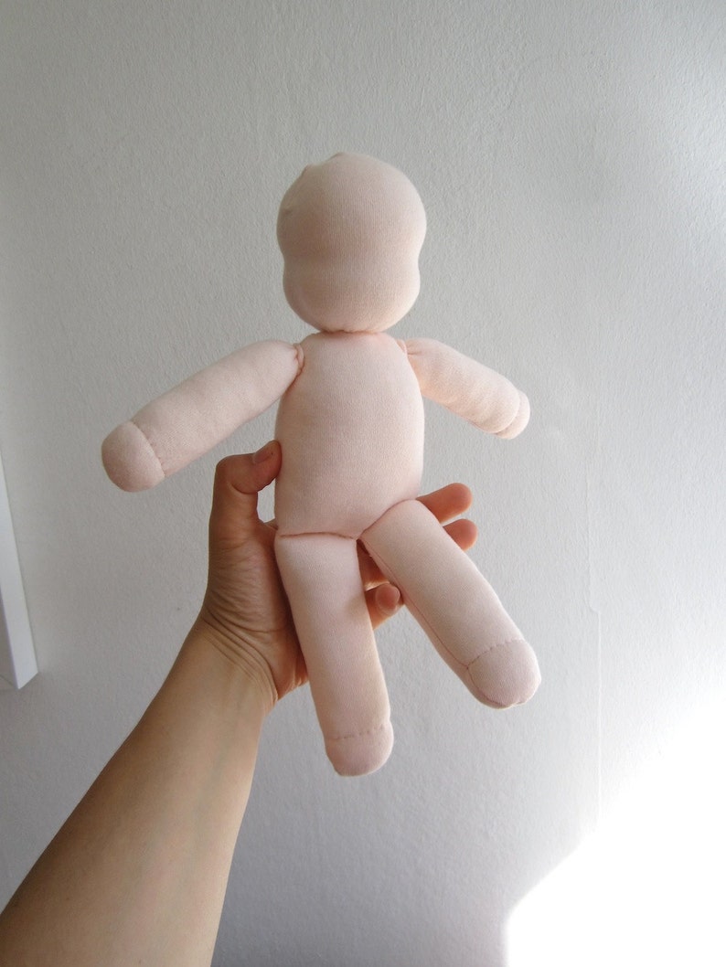 May include: A plain, light pink fabric doll with no facial features. The doll is held by a hand in the air.