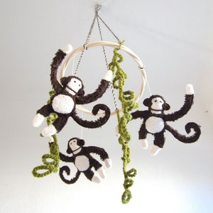 May include: A wooden ring mobile with three brown crocheted monkeys hanging from it. Each monkey has a white face and is holding onto a green crocheted vine. The mobile is suspended from the ceiling with three strings.