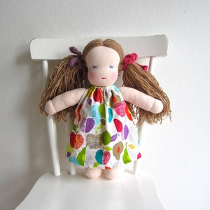 May include: A handmade doll with brown hair, blue eyes, and a colorful fruit-patterned dress. The doll is sitting on a white chair.