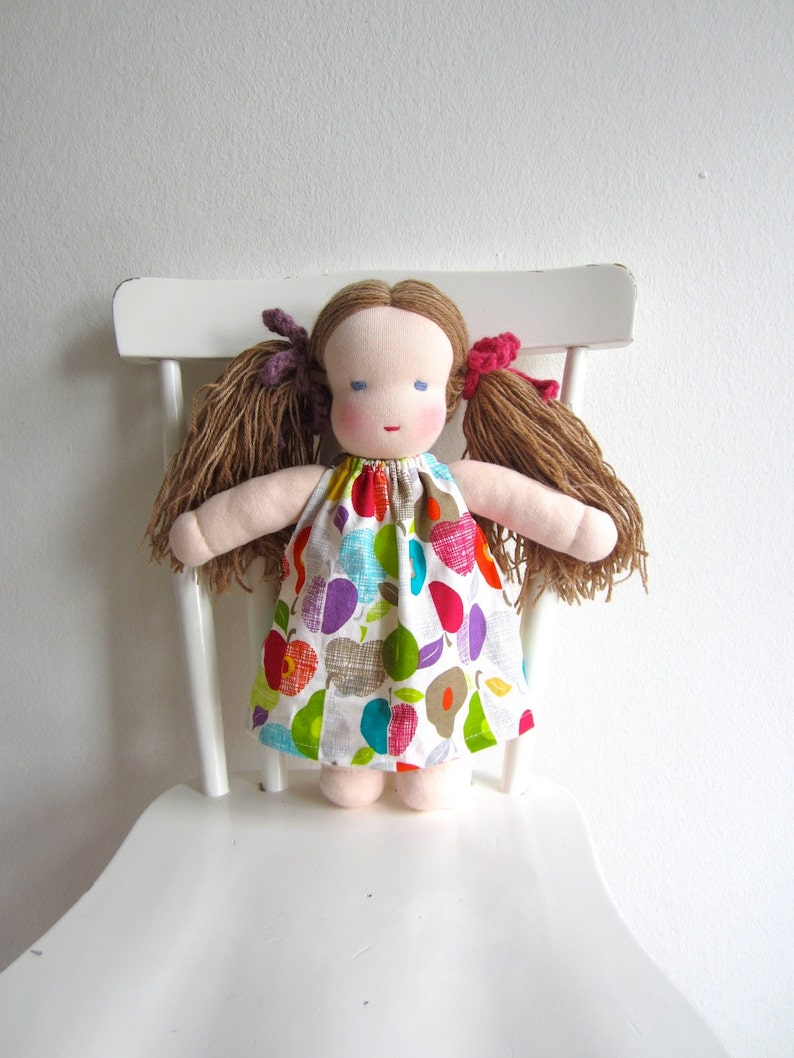 May include: A handmade doll with brown hair, blue eyes, and a colorful fruit-patterned dress. The doll is sitting on a white chair.