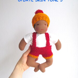 May include: A handmade doll with brown skin, wearing a white shirt, red knitted overalls with orange trim, and a yellow knitted hat with a red pom-pom. The doll is being held by a hand.