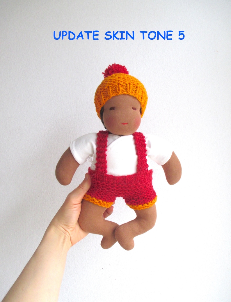 May include: A handmade doll with brown skin, wearing a white shirt, red knitted overalls with orange trim, and a yellow knitted hat with a red pom-pom. The doll is being held by a hand.