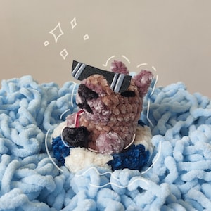 Puede incluir: Un oso de peluche hecho a mano con gafas de sol, flotando en una boya azul y blanca. El oso está rodeado por una manta esponjosa de color azul claro. El oso tiene una nariz negra y lleva gafas de sol negras con rayas blancas.