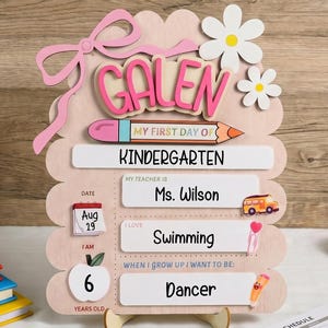Pode incluir: Uma placa de madeira para o primeiro dia de jardim de infância com o nome Galen em letras cor-de-rosa, um gráfico de lápis e painéis brancos com texto incluindo Ms. Wilson, Swimming e Dancer, decorada com um laço rosa e flores brancas.