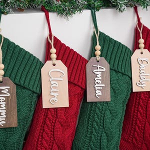 Puede incluir: Cuatro calcetines navideños de punto trenzado rojo y verde con etiquetas de nombre de madera. Las etiquetas dicen "Mommy", "Claire", "Amelia" y "Daddy". Cada etiqueta está adornada con cuentas de madera y colgada con hilo.
