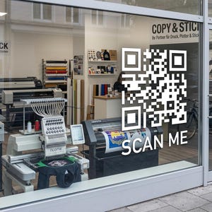 Könnte beinhalten: Ein großer weißer QR-Code-Aufkleber auf einer Schaufensterscheibe mit dem darunter gedruckten Text SCAN ME. Durch das Glas ist eine Ricoma-Stickmaschine sowie verschiedene Vinylrollen und Druckgeräte im Ladeninneren zu sehen.