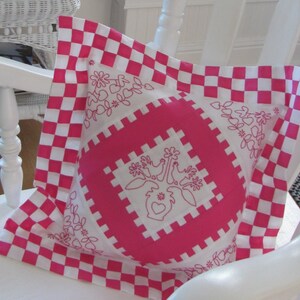 May include: A pink and white checkered pillow with embroidered white flowers and a heart design. The pillow is sitting on a white chair.