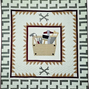 May include: A white quilt with a brown and red border. The center of the quilt features a brown toolbox with various tools inside. The toolbox is surrounded by a brown and white geometric pattern. The quilt is framed by a gray and white geometric border.