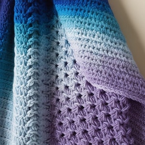 Könnte beinhalten: Eine gehäkelte Babydecke mit einem Farbverlauf von Blau, Türkis und Lila. Die Decke ist mit einem strukturierten Stichmuster gearbeitet.