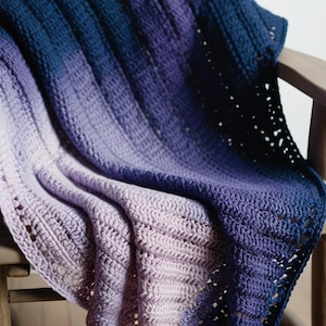 Op de afbeelding: Een gehaakt ombre deken in tinten paars en blauw, gedrapeerd over een houten stoel.