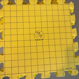 May include: Yellow interlocking foam mat with a grid pattern and the text "HEPHAESTUS CRAFTS". The mat has a puzzle-piece edge for connecting to other mats. The words "MADE IN CHINA" are printed on the mat.