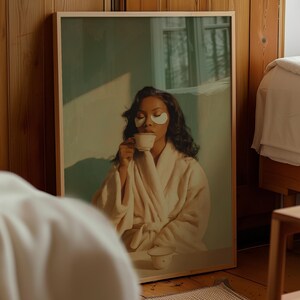 May include: A framed wall art print featuring a person with dark wavy hair wearing a beige robe and white under-eye patches. The person is holding a small white teacup to their lips, with a matching saucer resting on a table in the foreground.