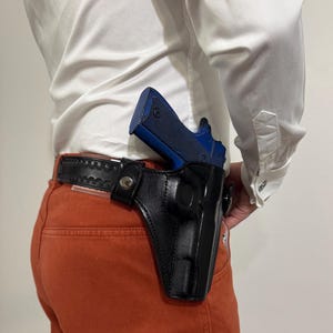 Könnte beinhalten: Ein schwarzes Lederholster, das an einem schwarzen Ledergürtel mit Ziernähten befestigt ist und eine blaue Handfeuerwaffe enthält. Das Holster verfügt über einen Metalldruckknopf und wird über einer orangefarbenen Hose und einem weißen Hemd getragen.