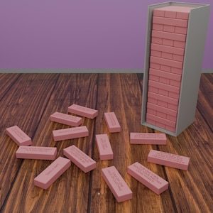 Könnte beinhalten: Ein rosa Blockstapelspiel mit Text auf den Blöcken. Einige Blöcke sind auf einer Holzoberfläche verstreut. Das Spiel beinhaltet einen Turm aus rosa Blöcken und einen grauen Behälter. Der Text auf den Blöcken enthält Sätze wie "Verführe deinen Partner mit Worten."