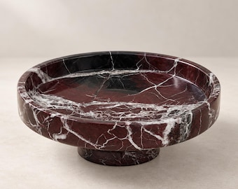 Puede incluir: Un cuenco circular con pedestal fabricado en mármol rojo burdeos intenso con vetas blancas prominentes en toda la superficie. El cuenco presenta una base ancha y poco profunda, junto con un soporte grueso y robusto de piedra pulida.