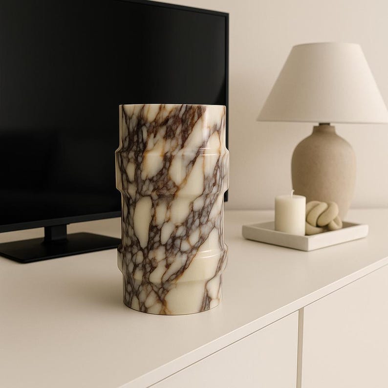 May include: A cylindrical vase with a cream and brown marble pattern. The vase has a textured surface and a geometric design. The vase is on a white surface, with a lamp and candles in the background.